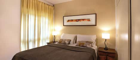 A neatly made bed with a dark bedspread, two bedside lamps, a ceiling fan, and a framed picture on the wall.