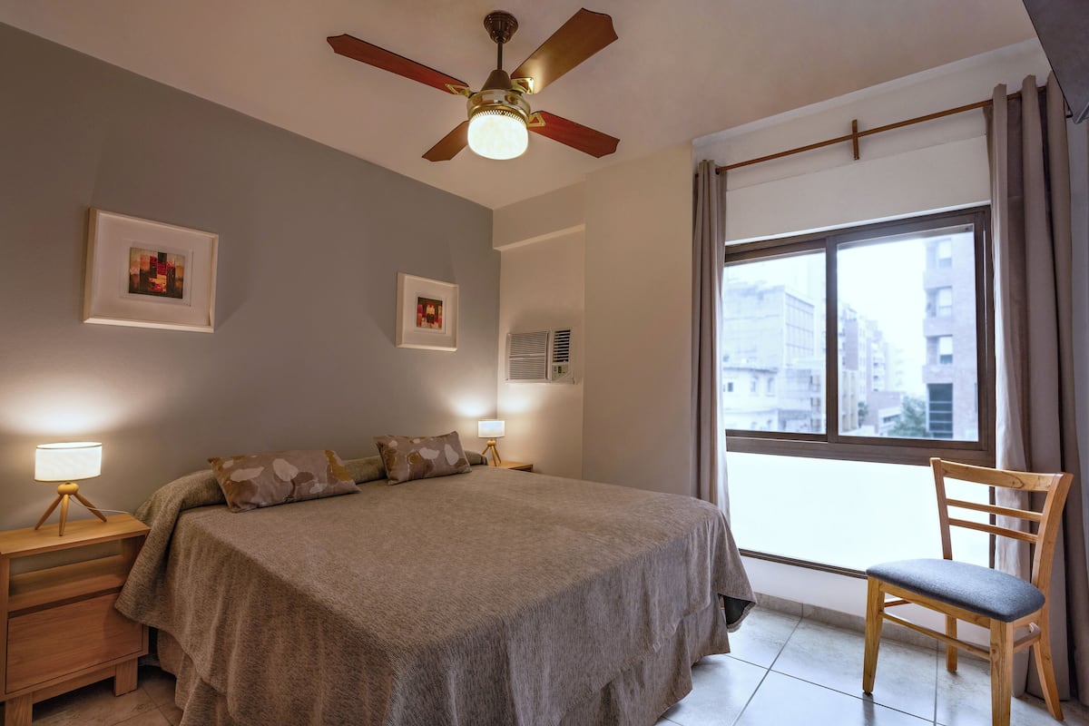 Un dormitorio con cama, una silla, ventilador de techo y ventana con cortinas.