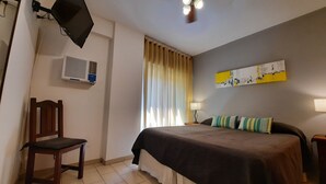 A hotel room with a bed, a chair, a TV mounted on the wall, an air conditioning unit, a window with curtains, and a painting on the wall. - Concord Rent Apart (Córdoba)