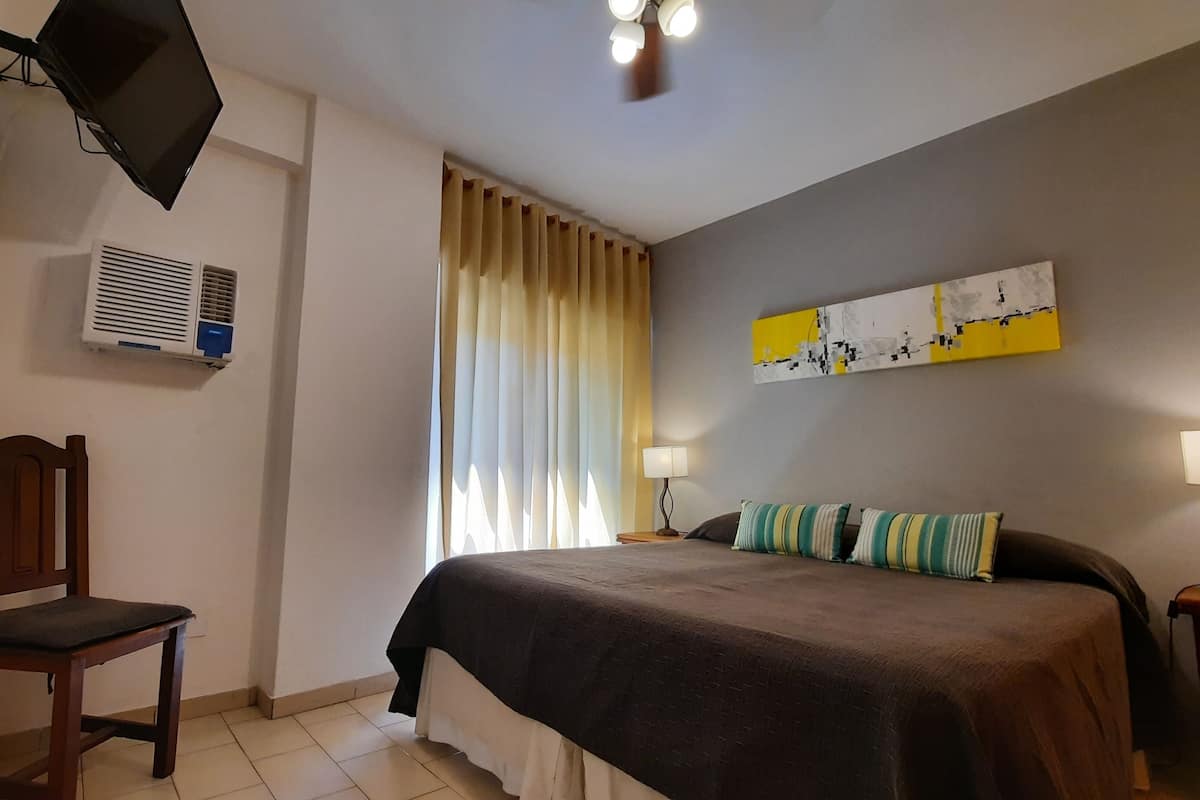 Habitación de hotel con cama, una silla, televisión montada en la pared, equipo de aire acondicionado, ventana con cortinas y un cuadro colgado.