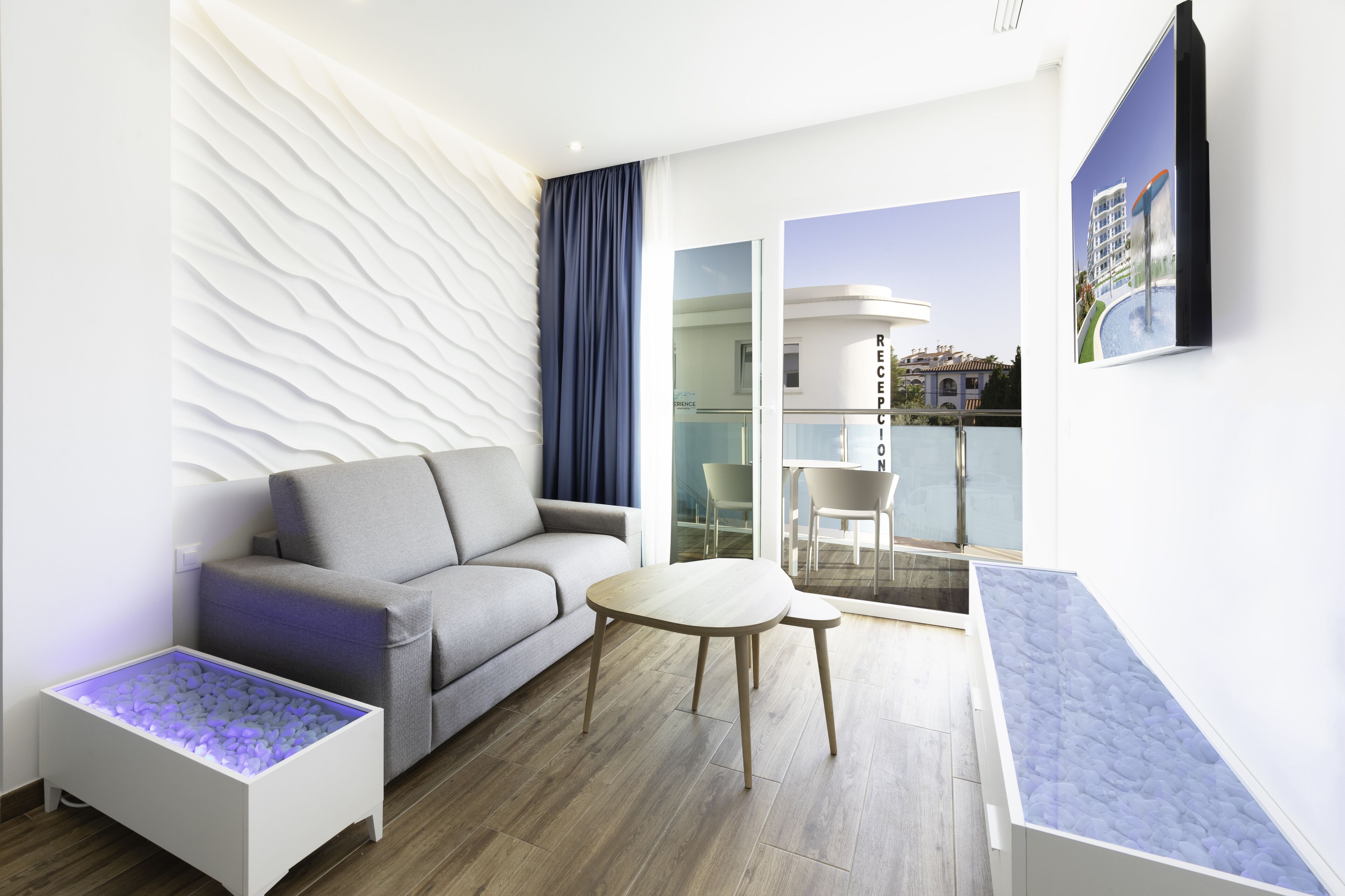 Een moderne hotelkamer met een grijze bank, een klein rond tafeltje en een witte bijzettafel met blauw verlichting.