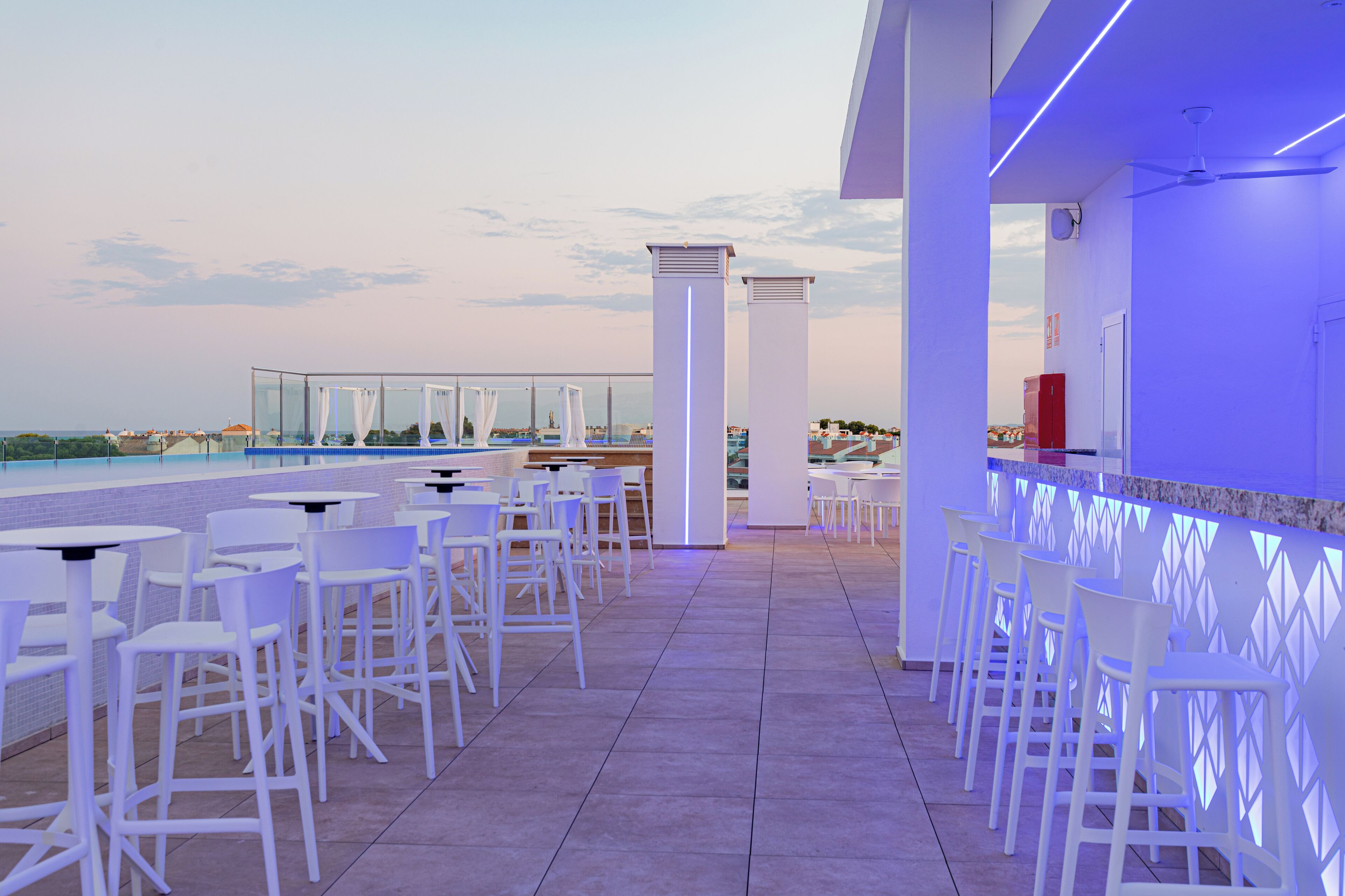 Un bar a la terrassa amb caires i taules blanques, una piscina i vista a l'horitzó de la ciutat.