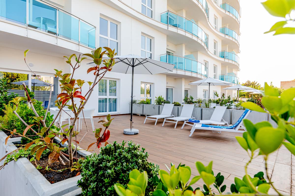 Terraza en la azotea con sillones de descanso y sombrillas, rodeada de vegetación y un edificio moderno con balcones.