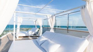 En balkon med seng og liggestole, der kigger ud over en strand og havet.