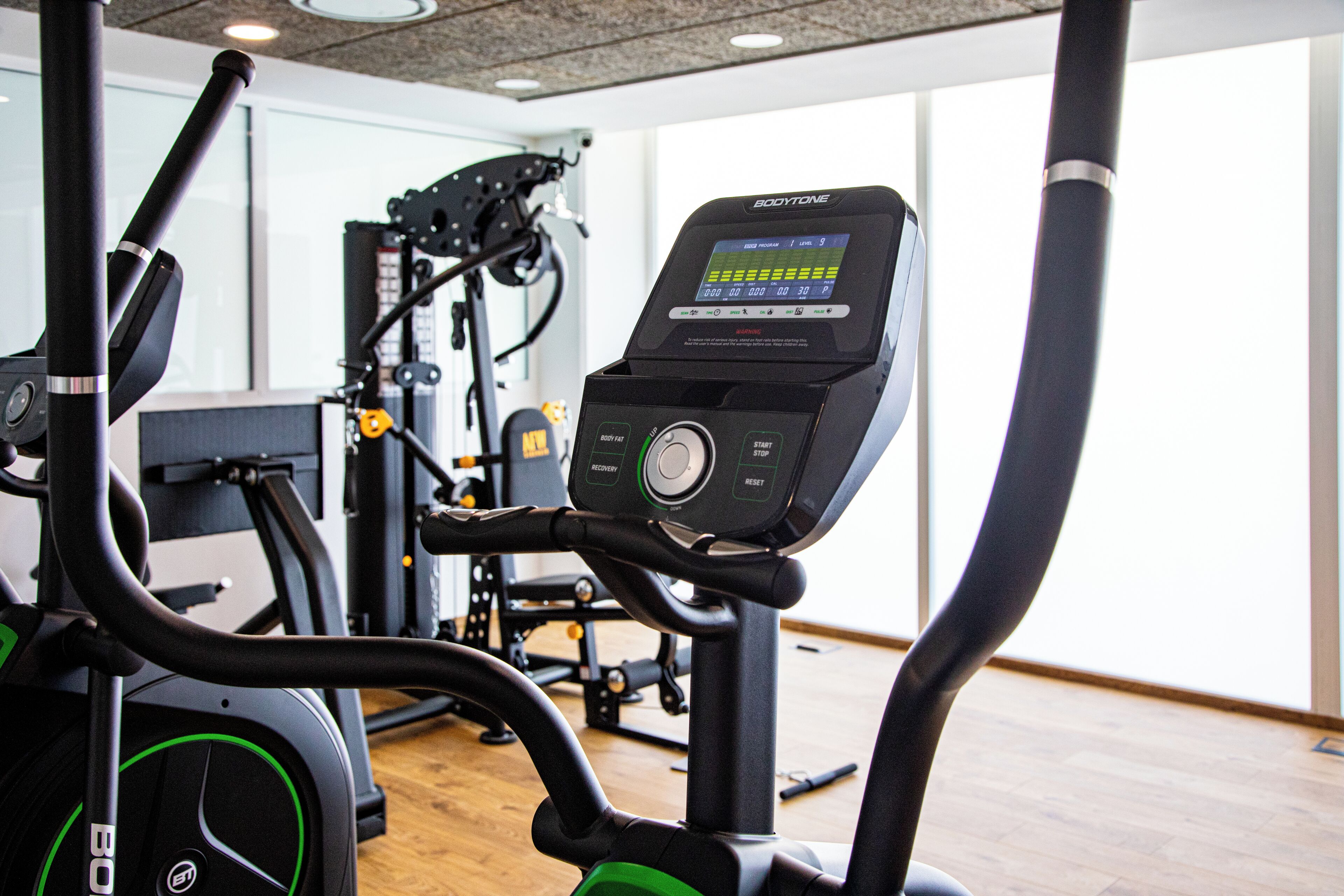 Una bicicleta estática con pantalla digital y panel de control, en una sala con equipo de ejercicio y ventanas amplias.