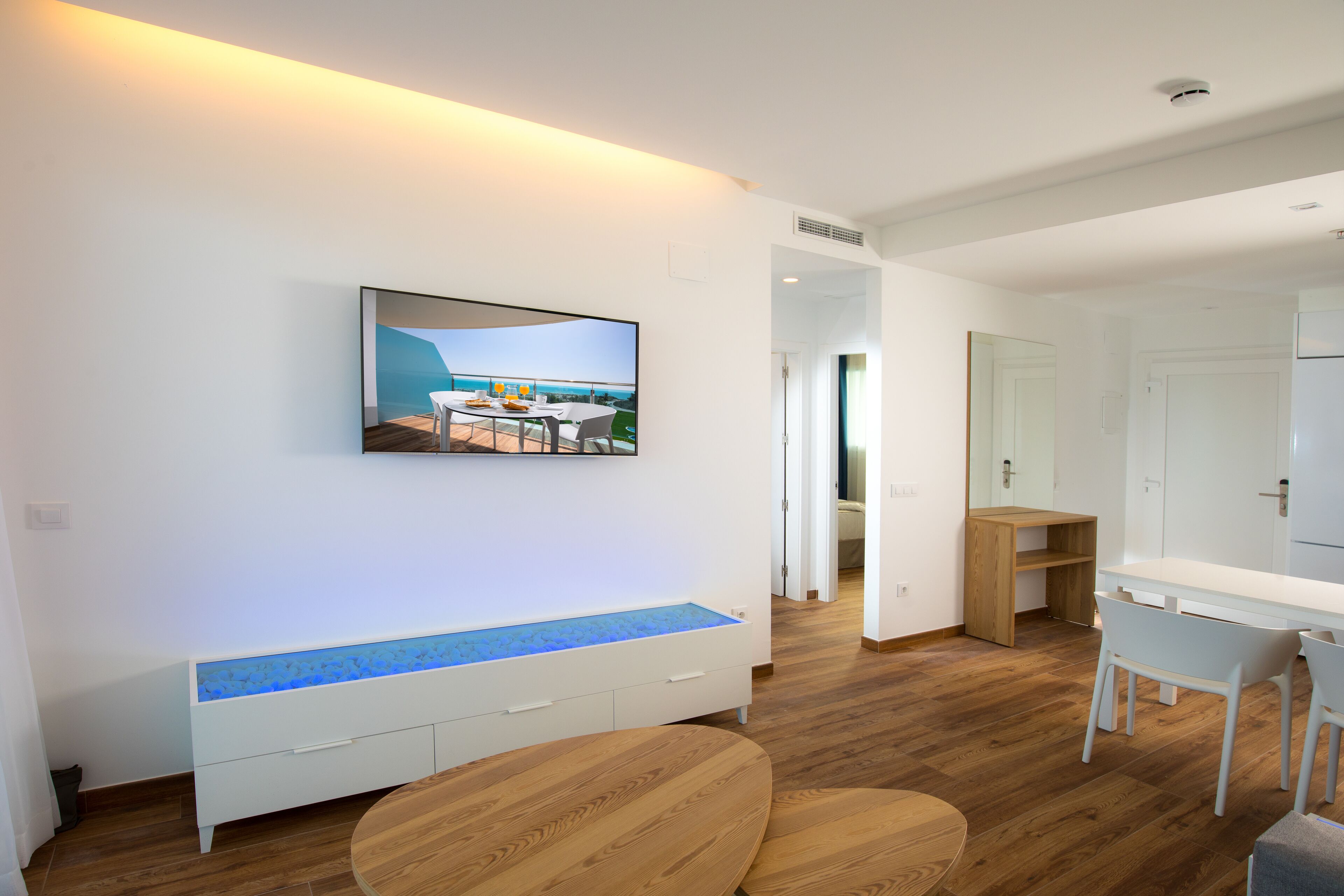 A modern living room with a flat-screen TV mounted on the wall, a wooden bench with a blue-lit edge, and a dining area with a table and chairs.