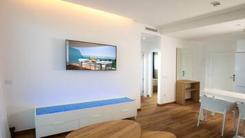 A modern living room with a flat-screen TV mounted on the wall, a wooden bench with a blue-lit edge, and a dining area with a table and chairs.