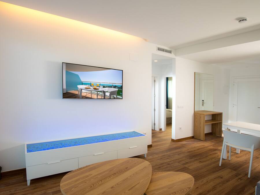 A modern living room with a flat-screen TV mounted on the wall, a wooden bench with a blue-lit edge, and a dining area with a table and chairs.