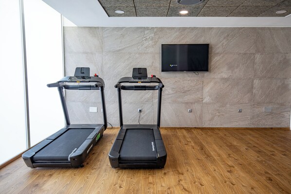 Dos cintas de correr en un gimnasio con suelo de madera, un televisor fijado a la pared y una ventana que deja entrar luz natural.