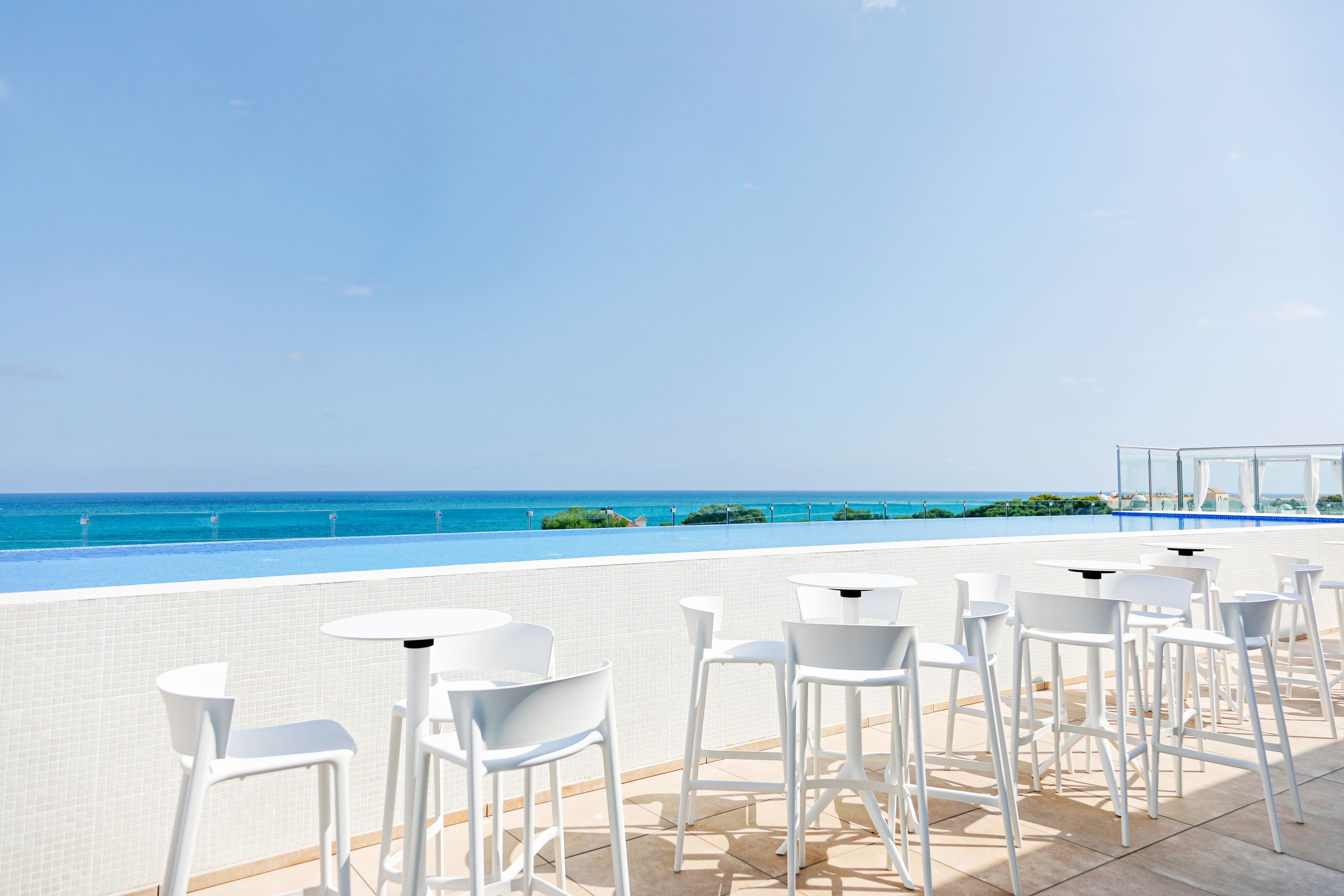 Terraza en la azotea con sillas y mesas blancas, con vista a una piscina y al océano.