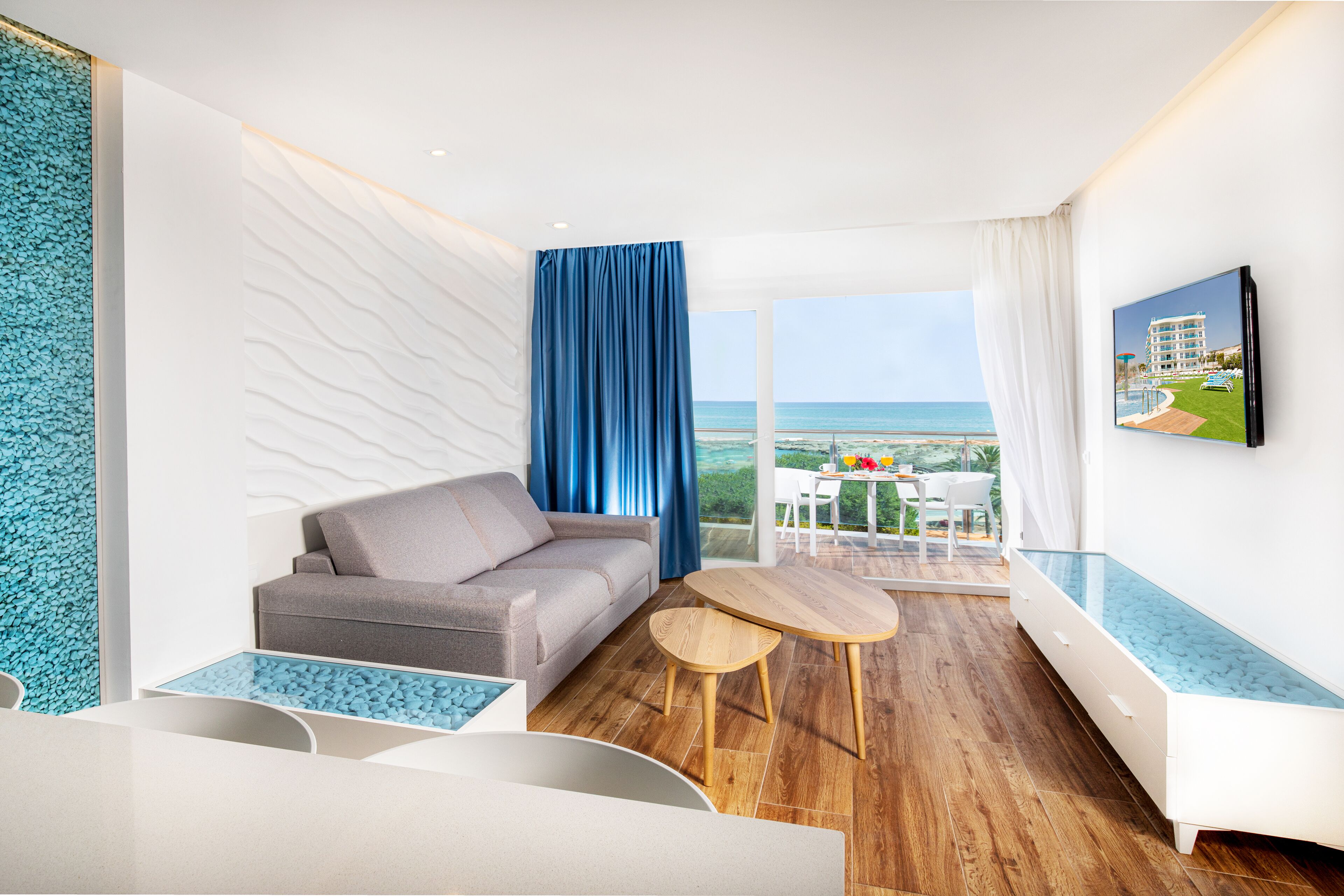 Une chambre d’hôtel moderne avec un canapé, une petite table, un téléviseur fixé au mur et un balcon offrant une vue sur la mer.