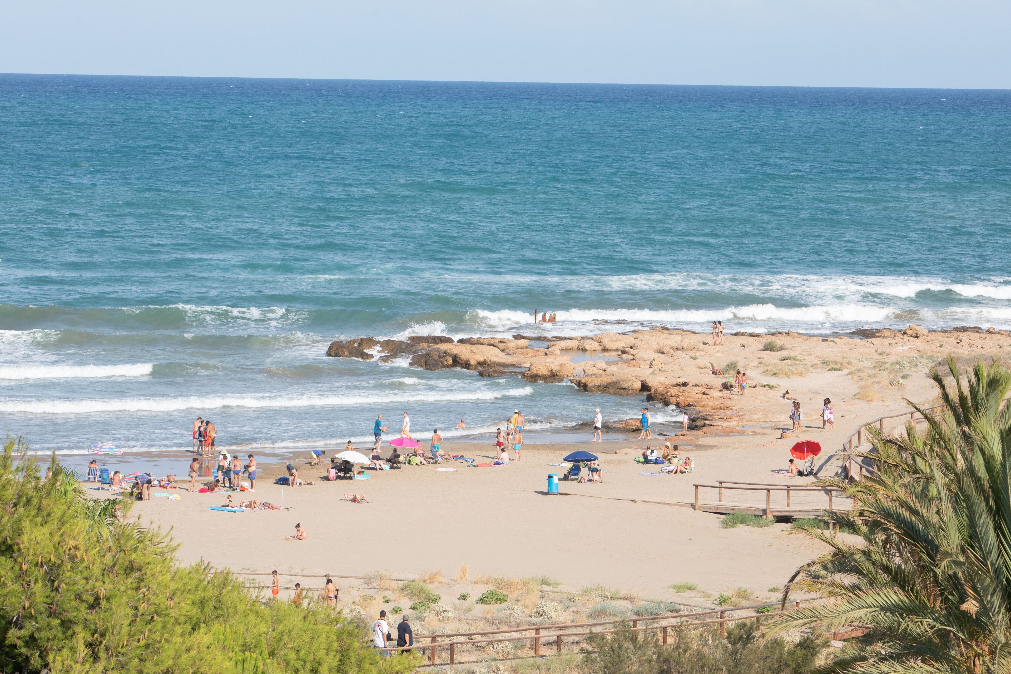 Una playa con gente relajándose, un pasillo de madera y formaciones rocosas cerca del agua.