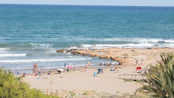Ein Strand mit entspannten Personen, einer Holzpromenade und Felsformationen am Wasser.