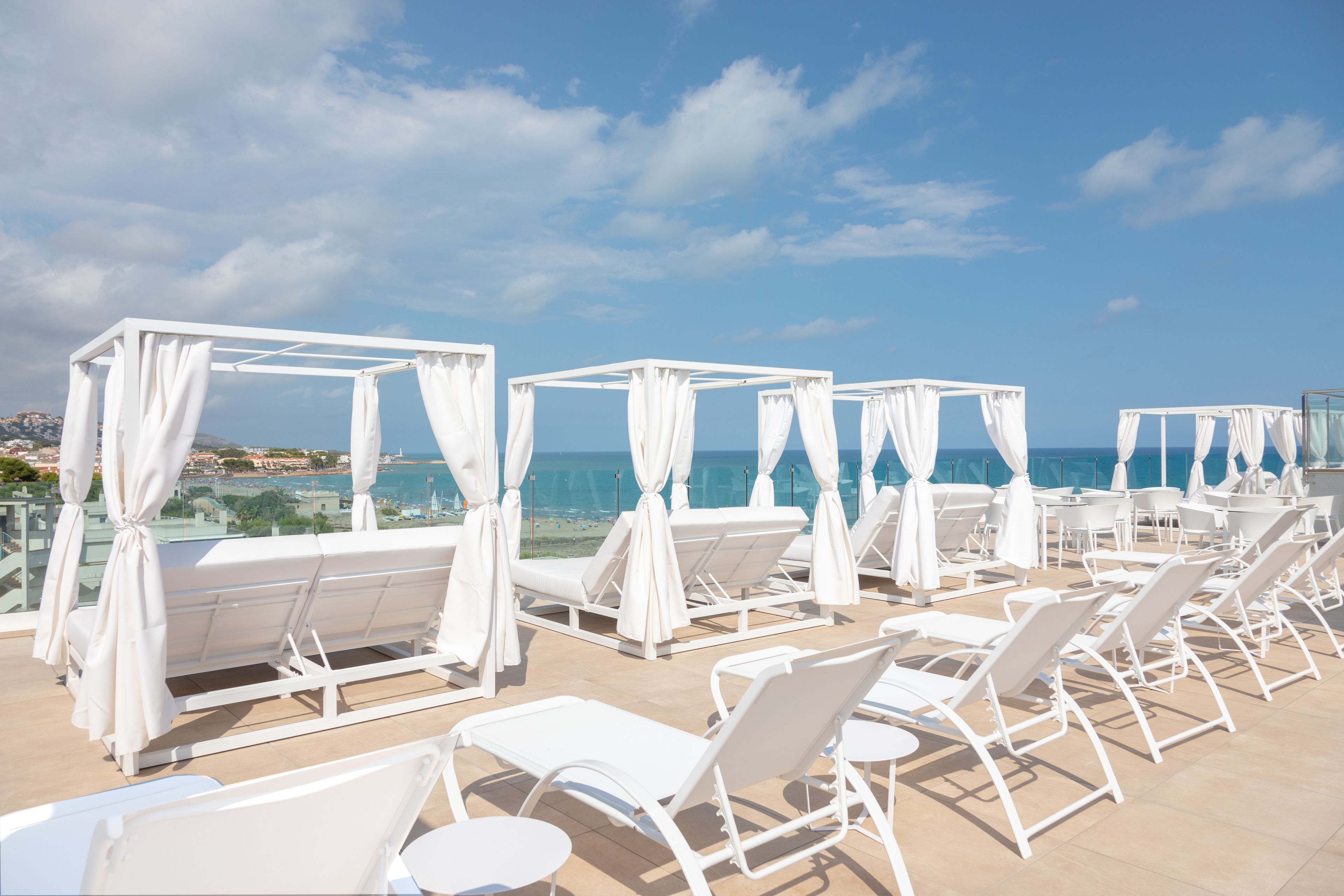A rooftop terrace with white sun loungers and canopies, overlooking a coastal view.