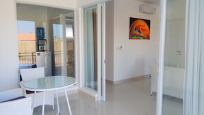 Quarto moderno com porta de vidro para uma varanda, uma mesinha redonda com cadeiras e um quadro abstrato colorido na parede.
