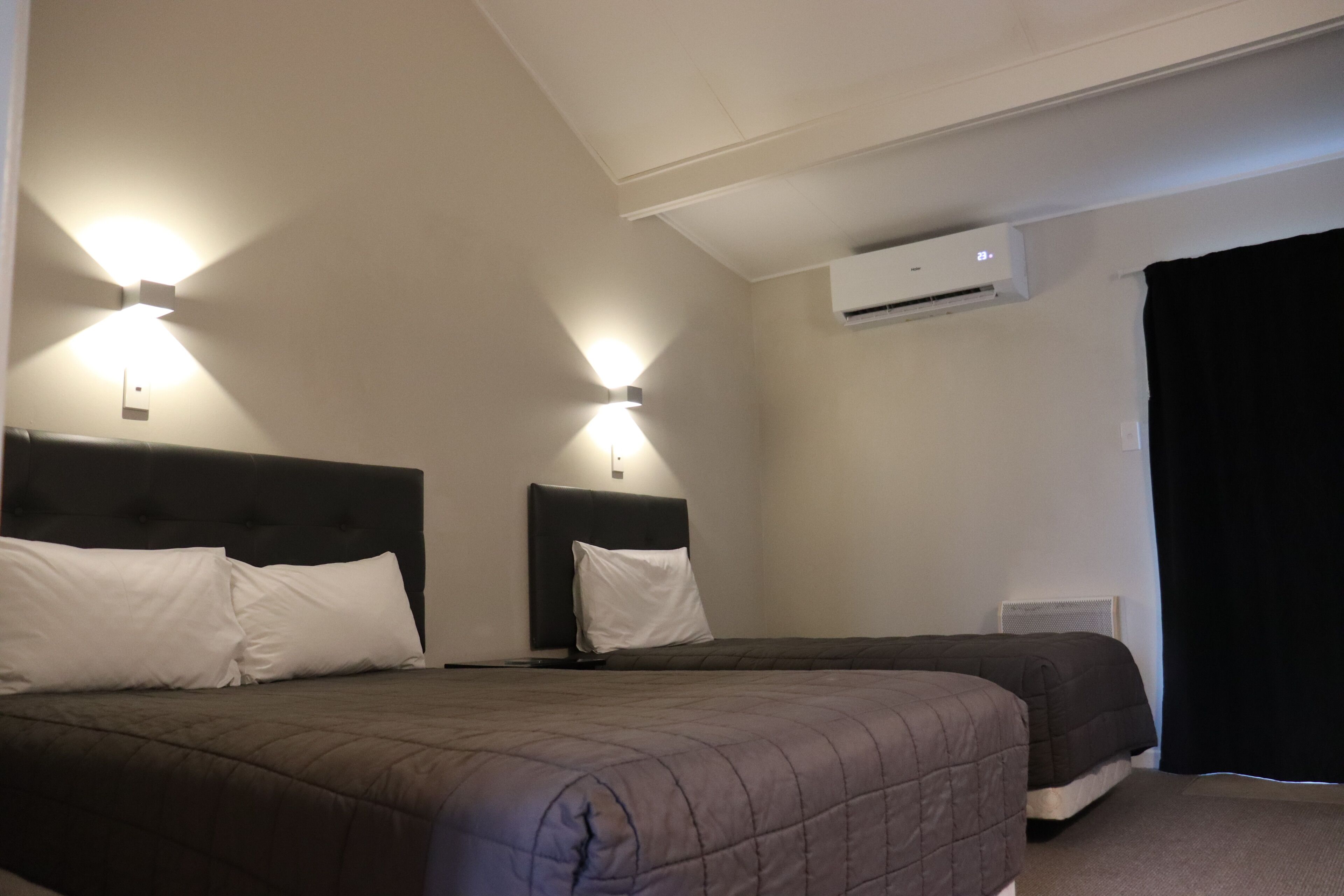 Et hotelværelse med to senge, en loftsvifter, en aircondition og et sort gardin.