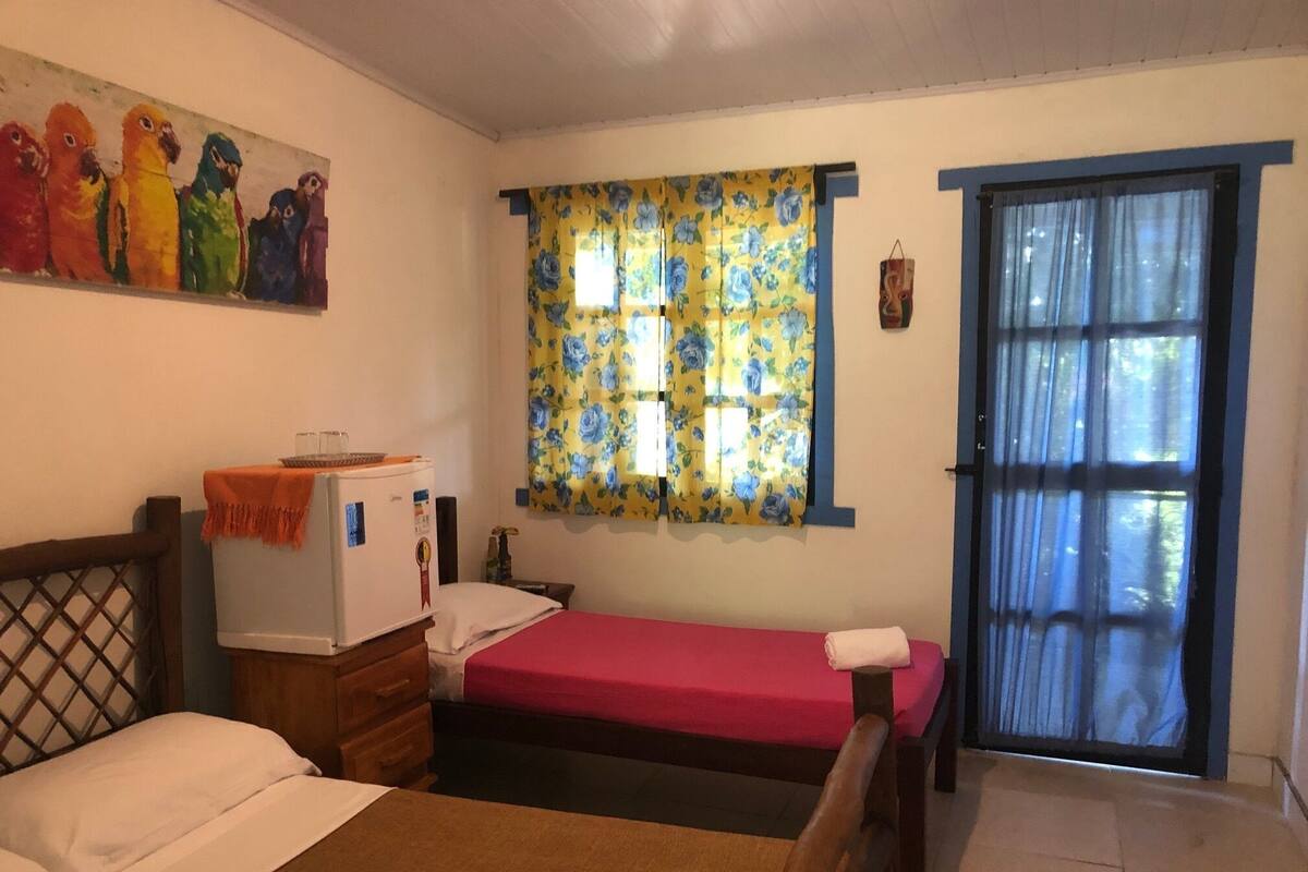 A room with two beds, a small refrigerator, a window with floral curtains, and a door with a blue frame.