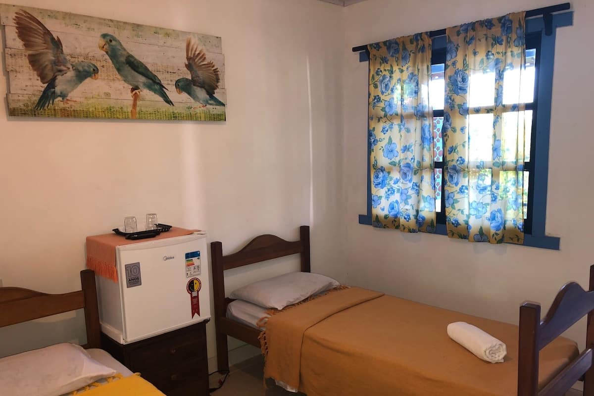 A small, simple room with two single beds, a small table with a refrigerator, a window with floral curtains, and a painting of birds on the wall.