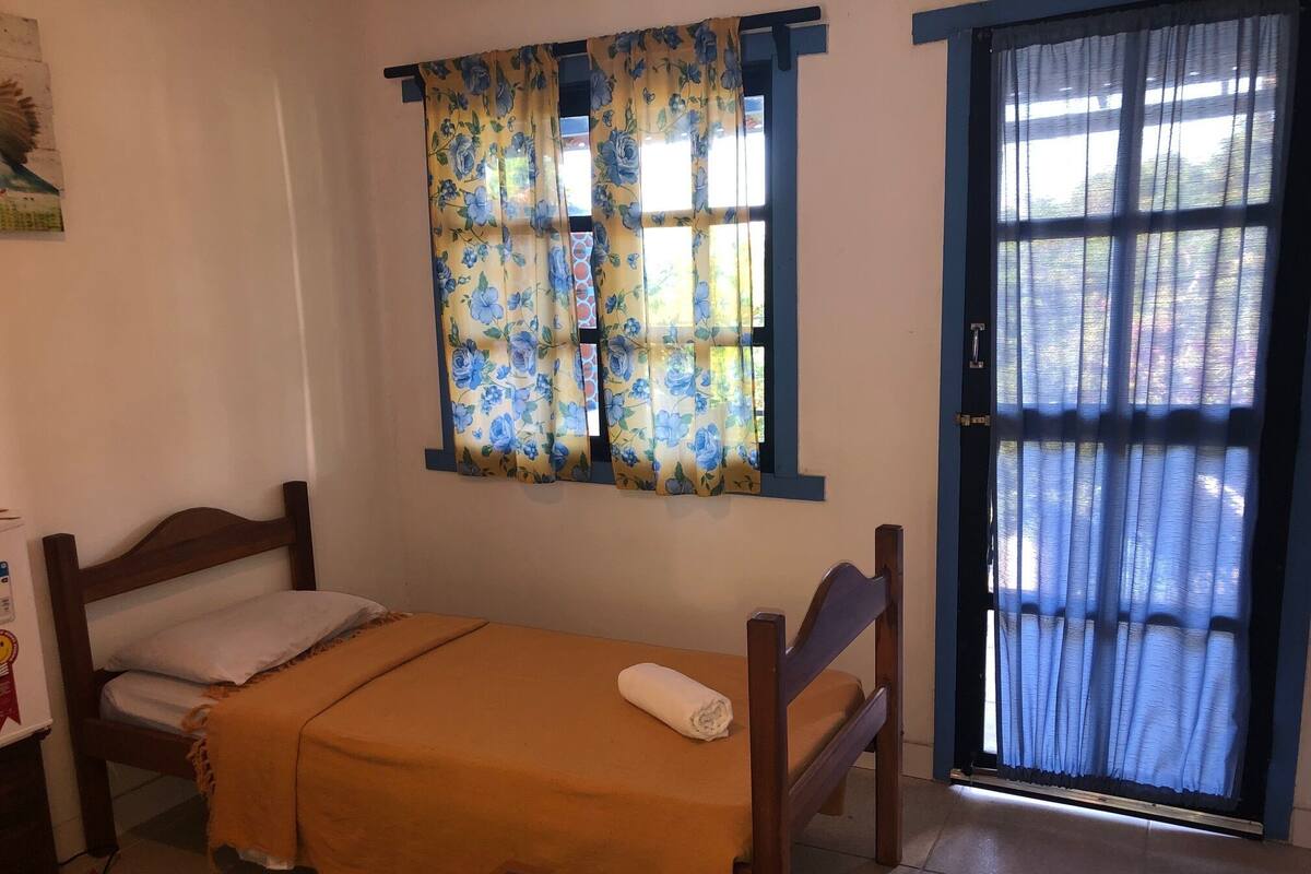 A small, single-bed room with a wooden headboard and footboard, a yellow bedspread, and a window with floral curtains.