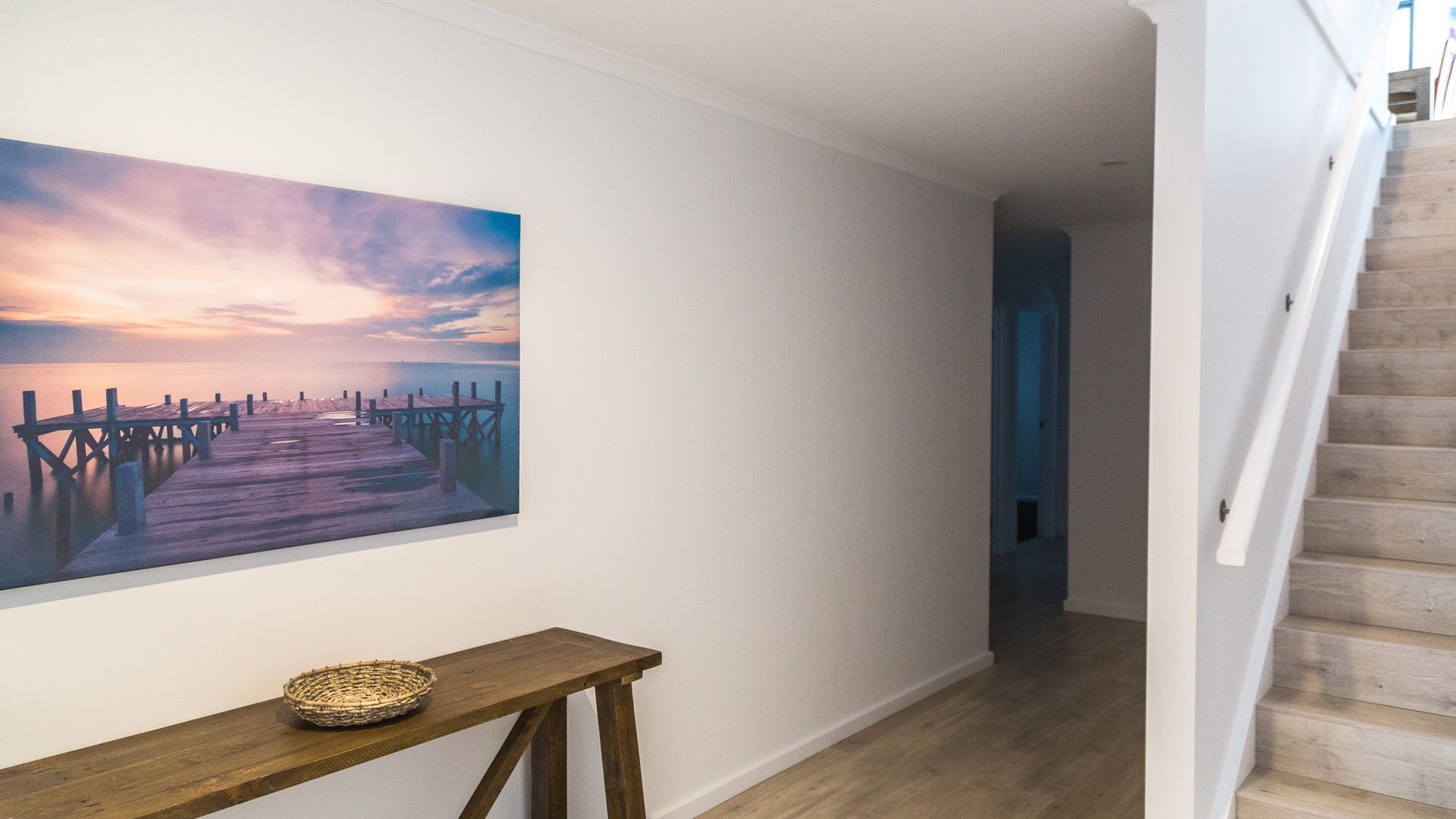 Un couloir avec un escalier en bois, un banc en bois surmonté d’un panier, et un grand tableau représentant une jetée au coucher du soleil.