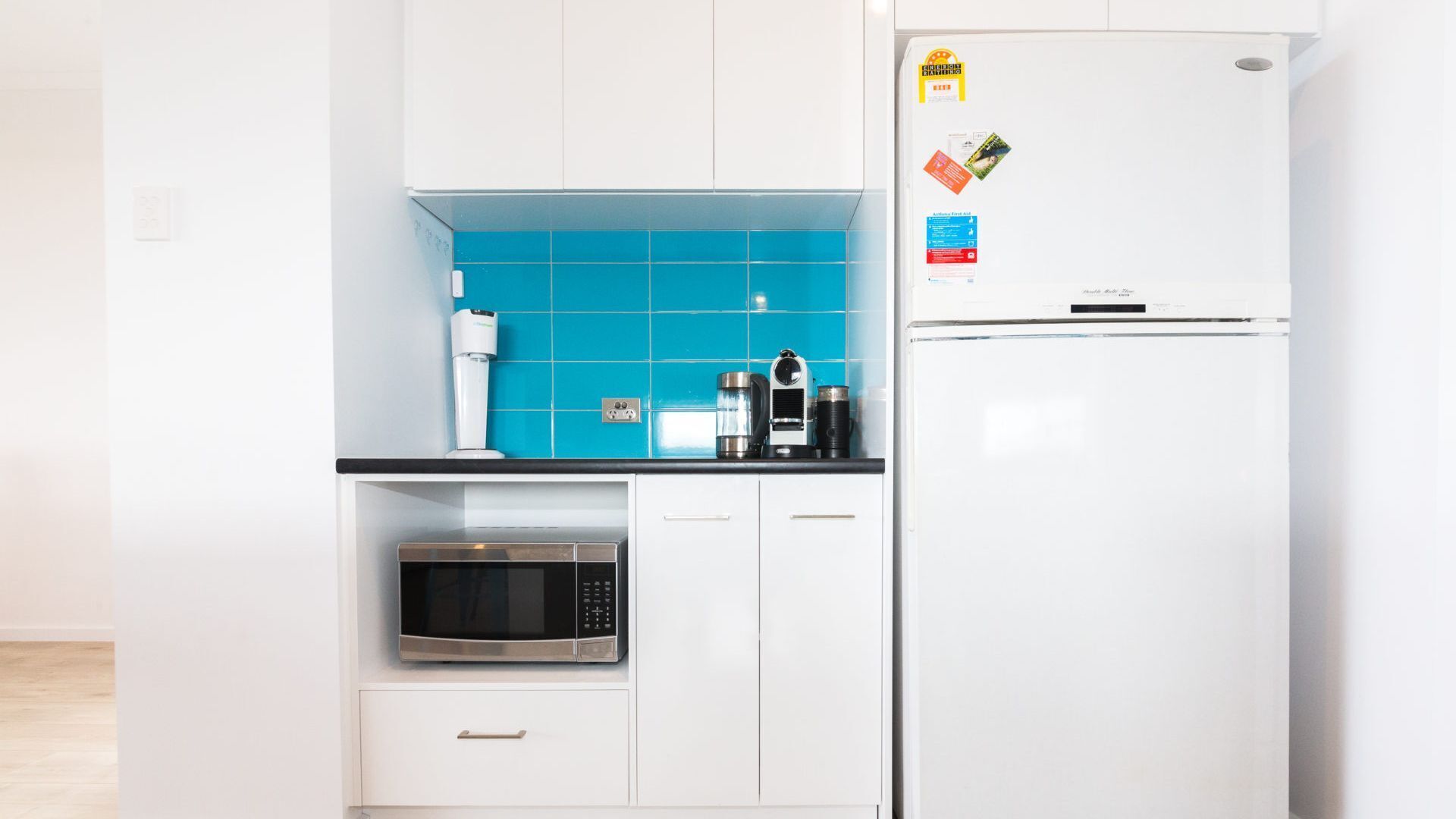 Une cuisine moderne avec des armoires blanches, un pare-feu bleu, un four à micro-ondes, une machine à café et un réfrigérateur décoré d’aimants.
