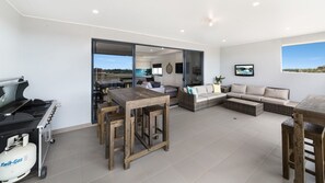 Een moderne keuken met een gasfornuis, een eethoek met een houten tafel en stoelen, een zithoek met een bank en een televisie, en uitzicht op de omgeving door een glazen deur.