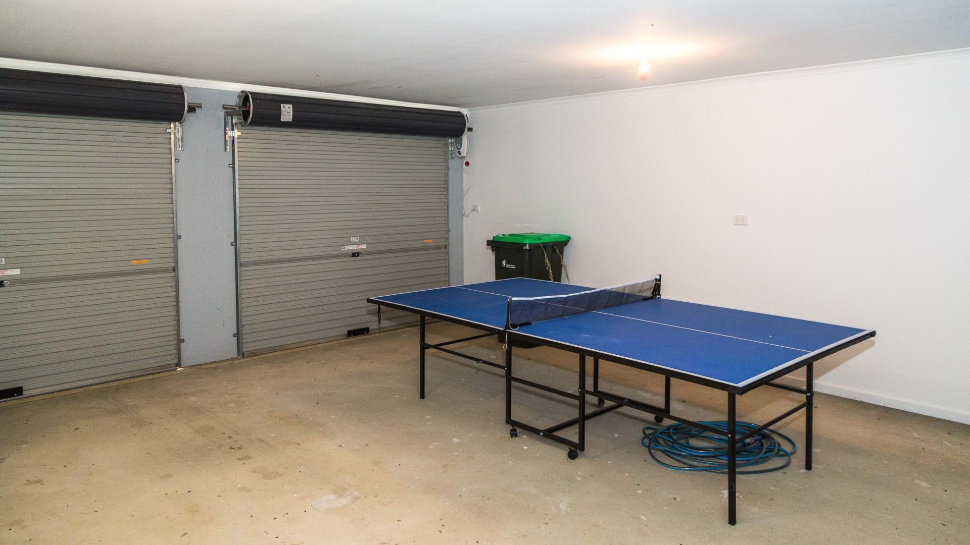 A ping pong table in a room with two closed garage doors and a green trash bin.