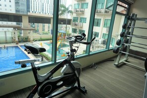 Espaço de ginástica interior com bicicleta ergométrica, suporte para pesos e vista para uma piscina e área de lazer ao ar livre.