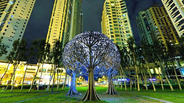 Vue nocturne d’un parc urbain moderne, avec une sculpture arborescente illuminée et originale au centre, entourée d’immeubles de grande hauteur.