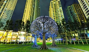A nighttime view of a modern urban park with a unique illuminated tree sculpture in the center, surrounded by high-rise buildings. - Pad in BGC Fort St.Luke (Taguig)