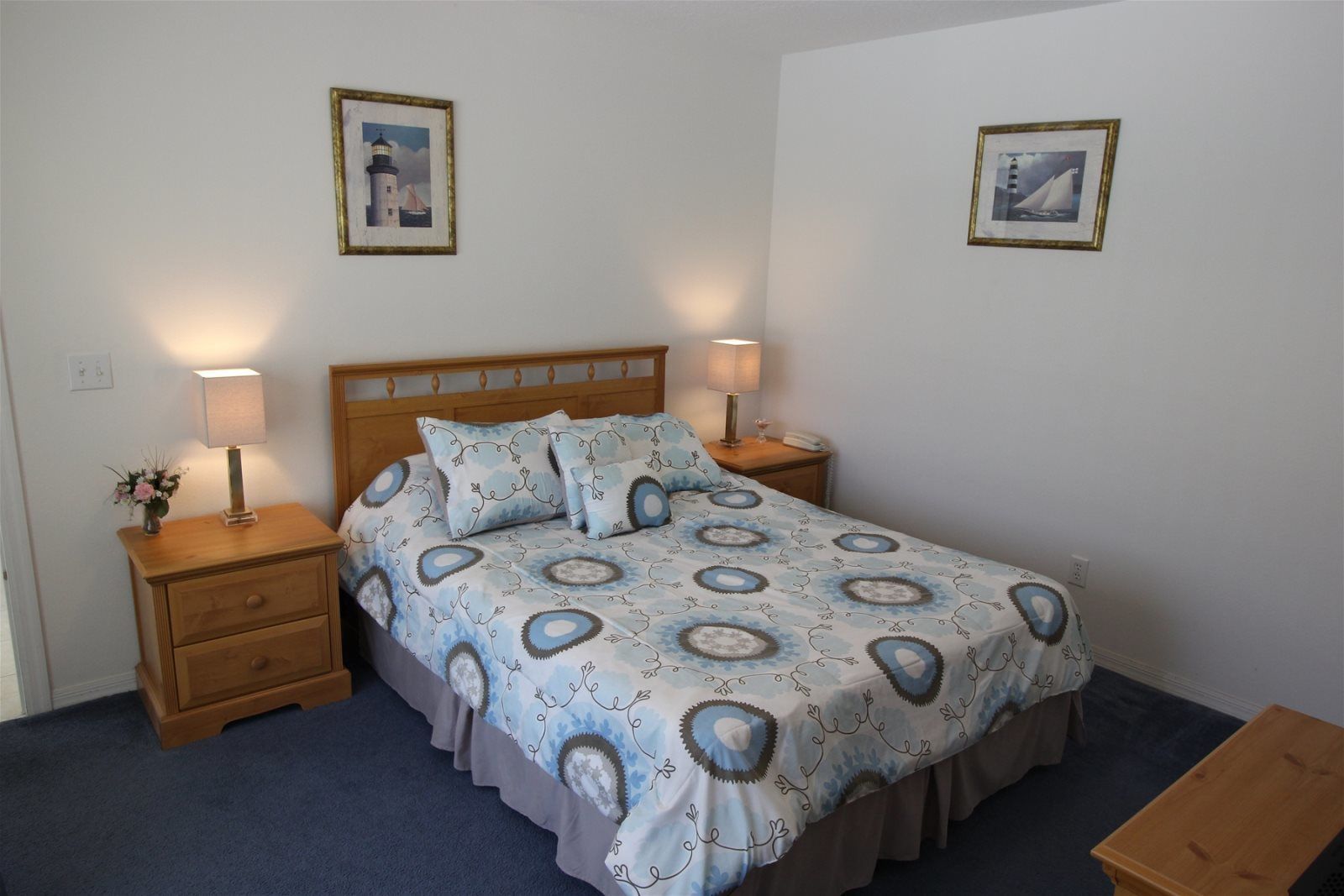 A neatly made bed with a patterned bedspread, two bedside lamps, a wooden nightstand with a vase of flowers, and two framed pictures on the wall.