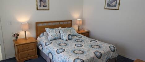 A neatly made bed with a patterned bedspread, two bedside lamps, a wooden nightstand with a vase of flowers, and two framed pictures on the wall.