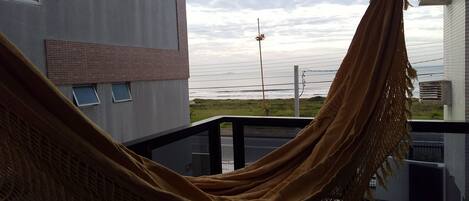 A hammock on a balcony overlooking a beach with buildings in the background.