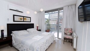Een slaapkamer met een groot bed, een aan de muur bevestigde televisie, een raam met gordijnen en een balkon met plantenpotten.