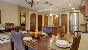 A modern kitchen with wooden cabinets, a stainless steel refrigerator, and a dining area with a wooden table set for four. - Aurora 225 | Resort Suite with Pool & Hot Tub (Canmore)