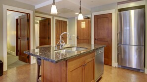 A kitchen with granite countertops, a stainless steel refrigerator, wooden cabinets, and pendant lighting. - Aurora 225 | Resort Suite with Pool & Hot Tub (Canmore)
