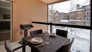 A balcony with a table set for two, a grill, and a view of snow-covered buildings. - Aurora 225 | Resort Suite with Pool & Hot Tub (Canmore)