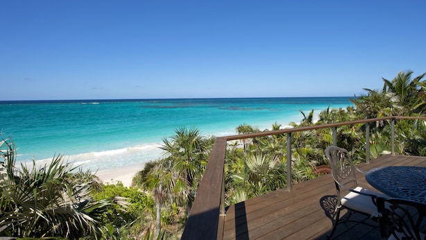 Eine Holzterrasse mit Tisch und Stühlen mit Blick auf einen Strand mit türkisfarbenem Wasser und Palmen.