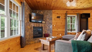 Interior acogedor de una cabaña con chimenea de piedra, un televisor montado encima y una zona de estar cómoda con sofá y mesa.