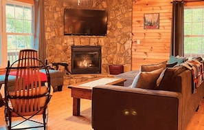 Interior acogedor de una cabaña con chimenea de piedra, un televisor de pantalla plana, un sofá de madera y un comedor con una silla.
