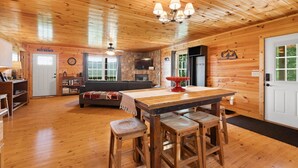 Interior de una cabaña de madera con una mesa de comedor, taburetes de bar, una chimenea de piedra y una zona de estar acogedora con un sofá y un televisor.