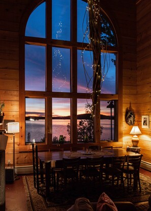 Une pièce chaleureuse avec une grande fenêtre en arc d’où l’on peut admirer un lac au coucher du soleil, des murs en bois, une table à manger avec des chaises et une lampe sur une table d’appoint.
