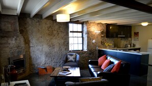 A rustic living room with a fireplace, a sofa, a coffee table, and a kitchen area with dark cabinets and a white refrigerator. - Heritage Garden Cottage (Hobart)