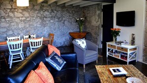 A cozy living room with a stone wall, a black leather sofa, a patterned armchair, a wooden coffee table, and a white side table with books and a vase of flowers. - Heritage Garden Cottage (Hobart)