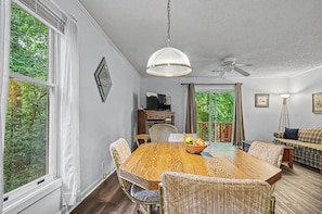 Un comedor con mesa de madera, sillas, una lámpara colgante y vistas al exterior con vegetación.