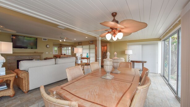 A spacious living room with a large wooden dining table, comfortable seating, a ceiling fan, and a television mounted on the wall.