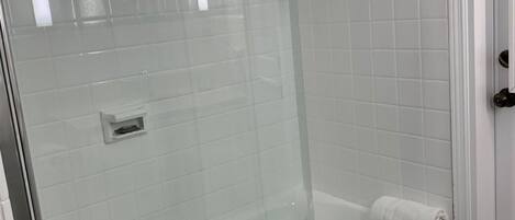 Un baño moderno con ducha de vidrio, una bañera blanca y un muro de azulejos con toallas colgadas en un perchero.
