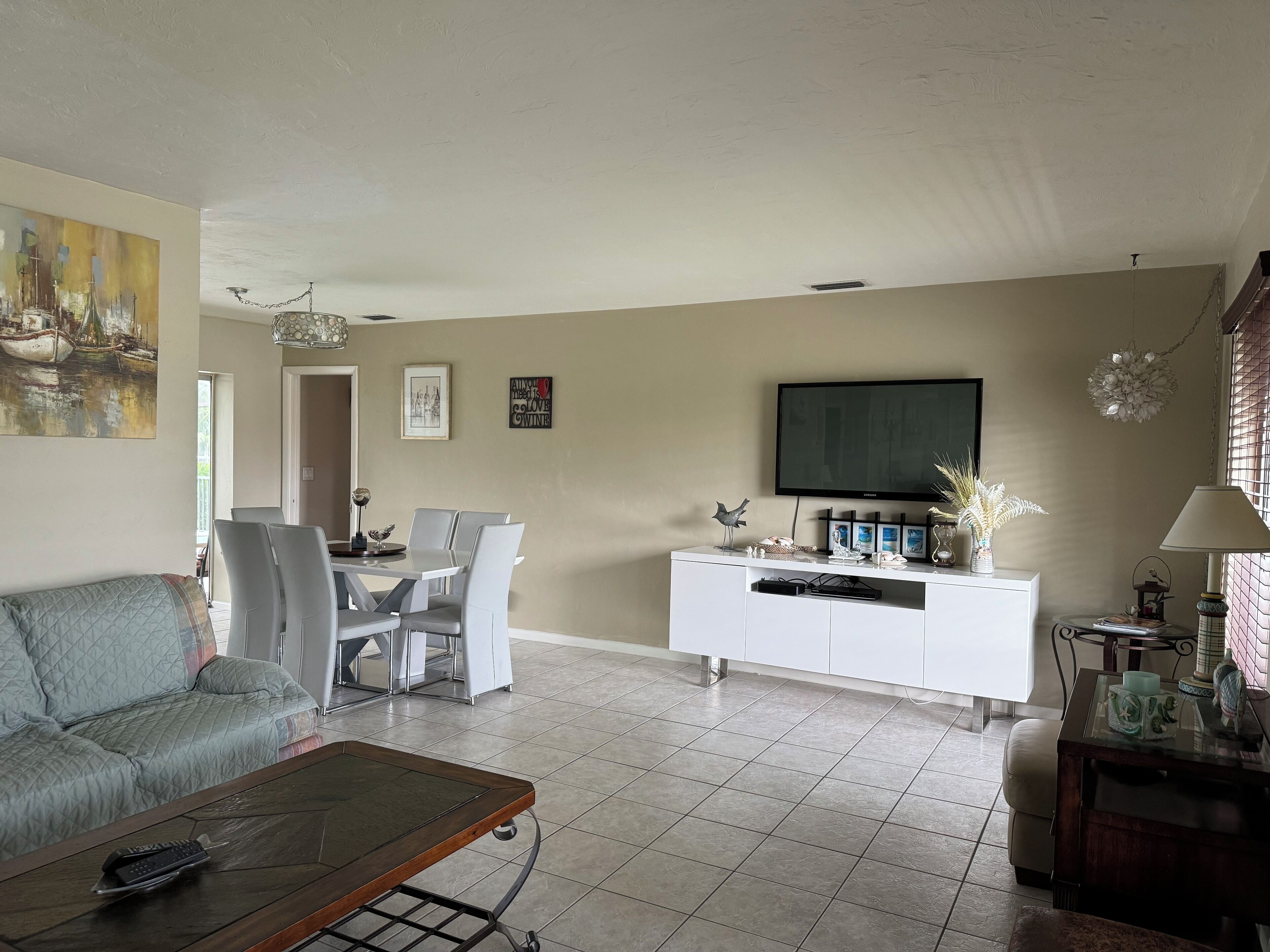 Uma sala de estar com sofá, mesa de centro, área de jantar com cadeiras, armário branco com objetos decorativos e TV fixada na parede.