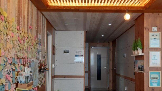 Un couloir avec des lambris en bois au plafond, un comptoir d’accueil où sont affichées des notes, et un espace détente avec des chaises.