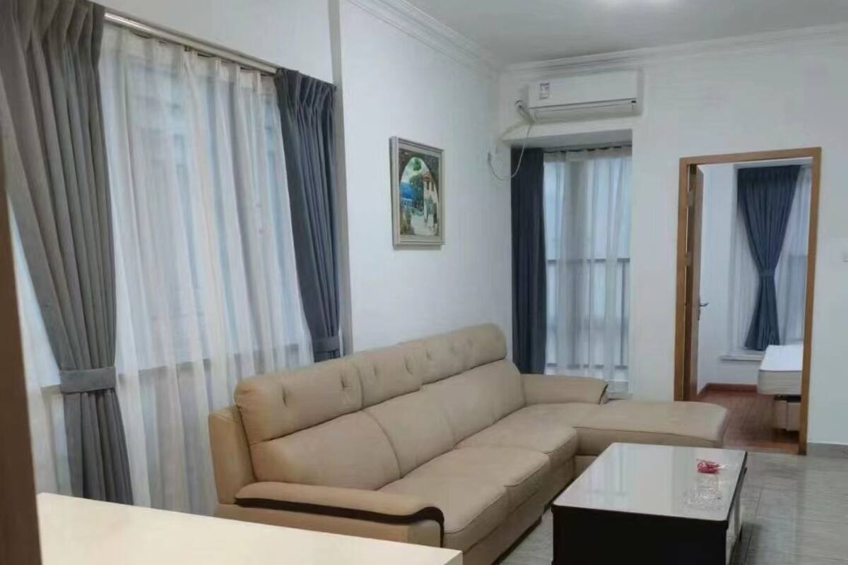Ein Wohnzimmer mit beiger Couch, Couchtisch und Fernseher. Vor den Fenstern hängen Vorhänge und an der Wand ist eine Klimaanlage zu sehen.